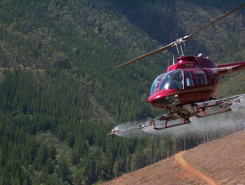 Red helicopter flying over a red-earth pasture while spraying chemicals. 