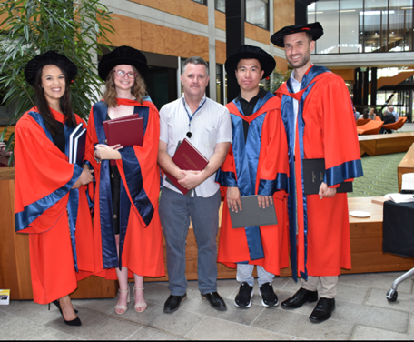 Five PhD graduates, four of them in red-and-blue gowns.