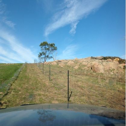 A fence in a paddock with rocks on one side and blue sky in the background.