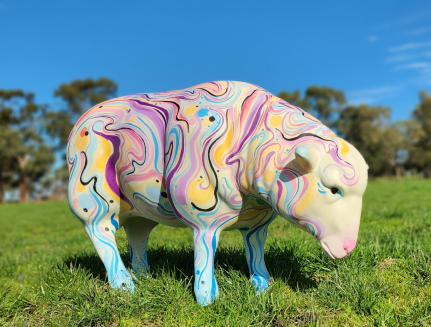 Sheep sculpture in a green, grassy paddock with rows of tall trees in the background. Sculpture is painted in contemporary style using a variety of colours, including: blue, purple, yellow and pink.  