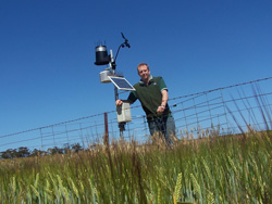 A man standing in a crop with weather station equipment, wire fence behind him