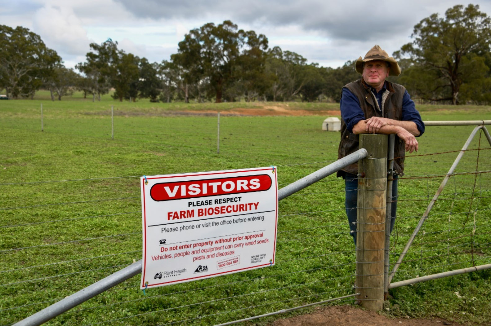 Farmer leaning on cattle gate next to sign which says 'Visitors, please respect Farm Biosecurity. Please phone or visit the office before entering. Do not enter property without prior approval. Vehicles, people and equipment can carry weed seeds, pests and diseases.