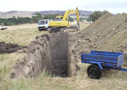 Trailer, truck and excavator next to a narrow, deep trench with vertical sides