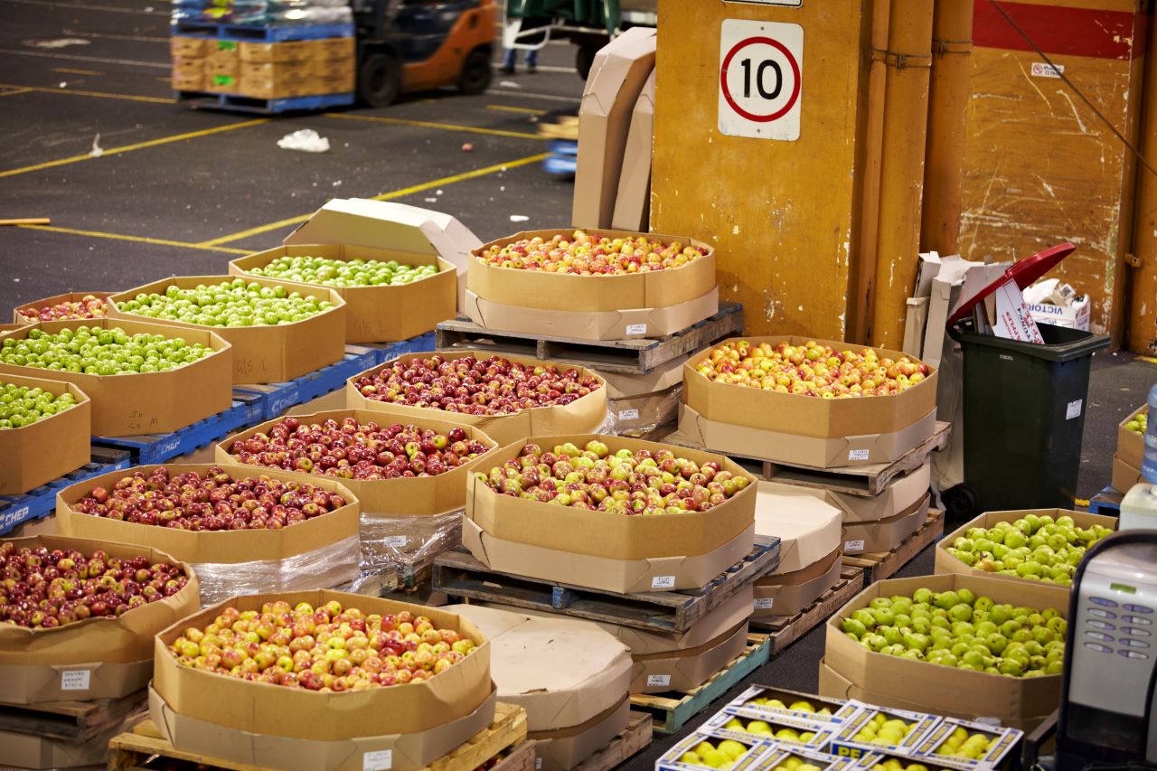 The image shows fruit bins at a market. There are various bins displaying red, yellow and green apples.