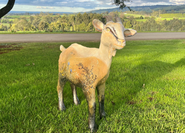 Goat sculpture standing on green grass with three goats and two people walking away in the background. Sculpture is painted in earthy tones with hand painted grass and flowers on the body of the goat. 