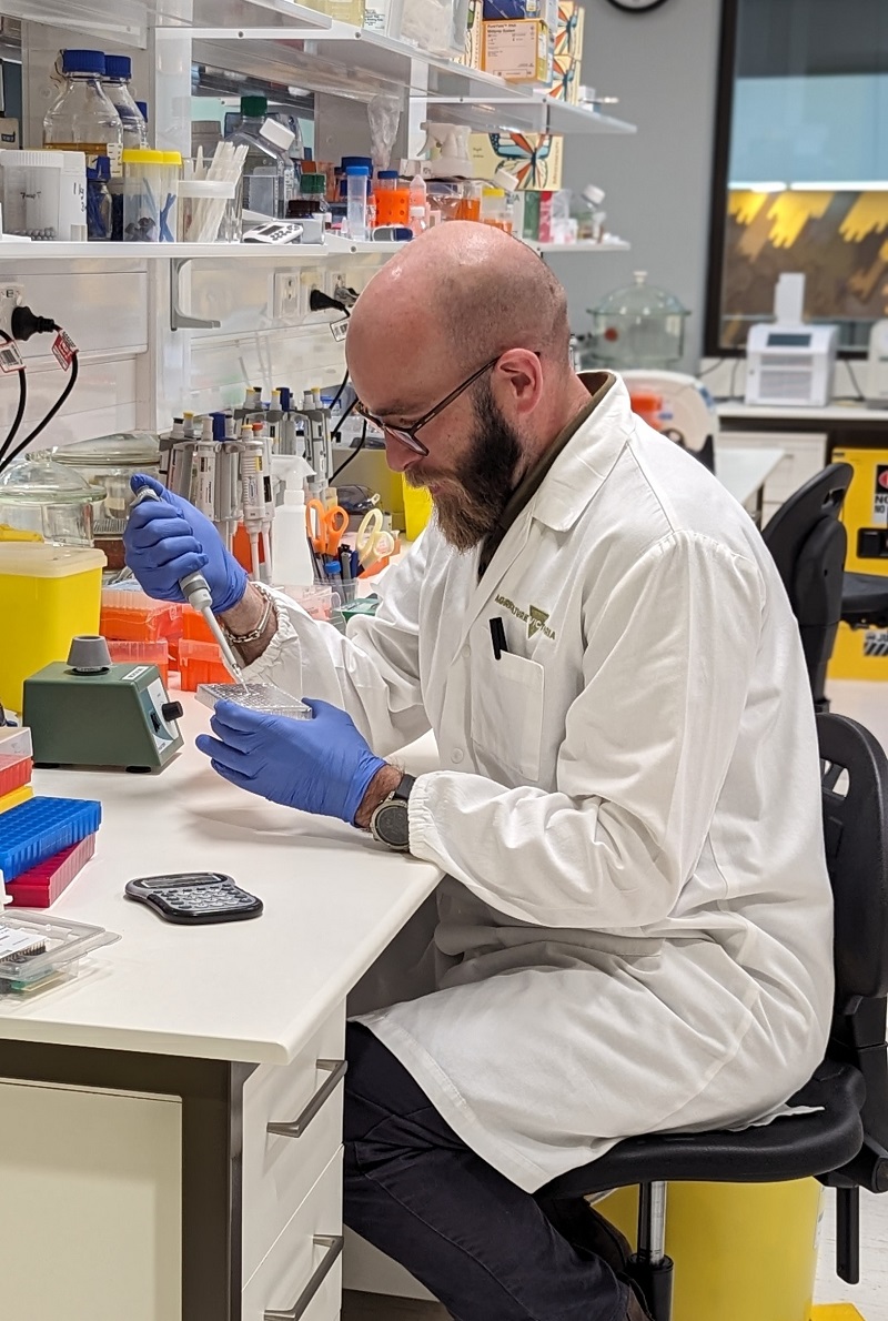 man in white lab coat wearing blue gloves holds eye dropper and petri dish while seated