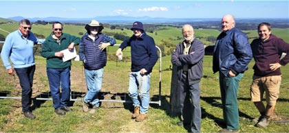 Seven farmers lined up against a fence on a hill, green valley in the background